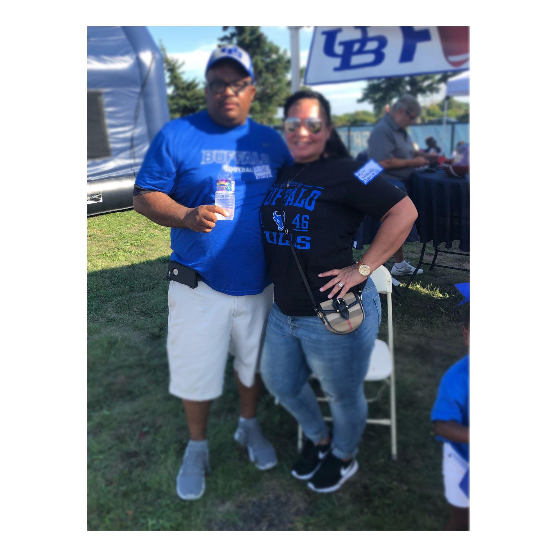 Two people wearing Buffalo Bulls shirts pose together outdoors, with a tent and people in the background.